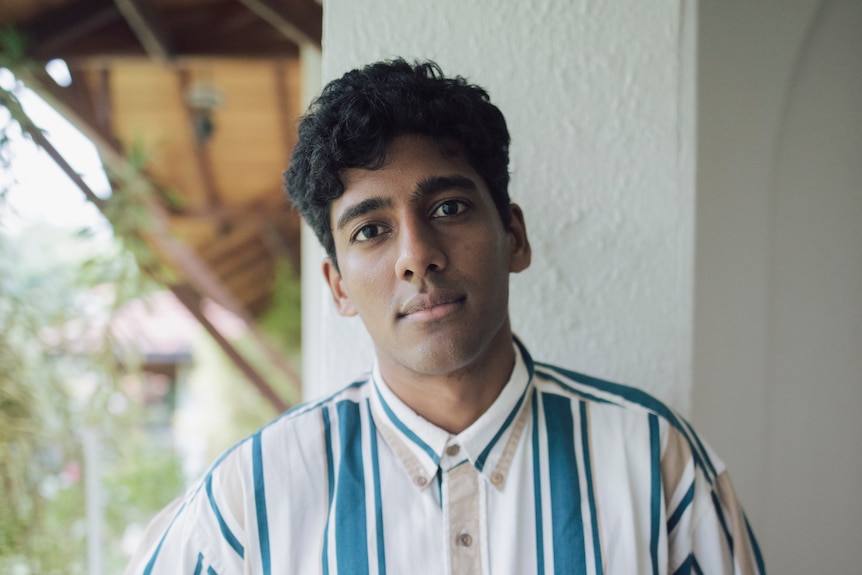 A young Sri Lankan man with short hair wears a blue and white striped shirt and stands on a veranda, looking at the camera.
