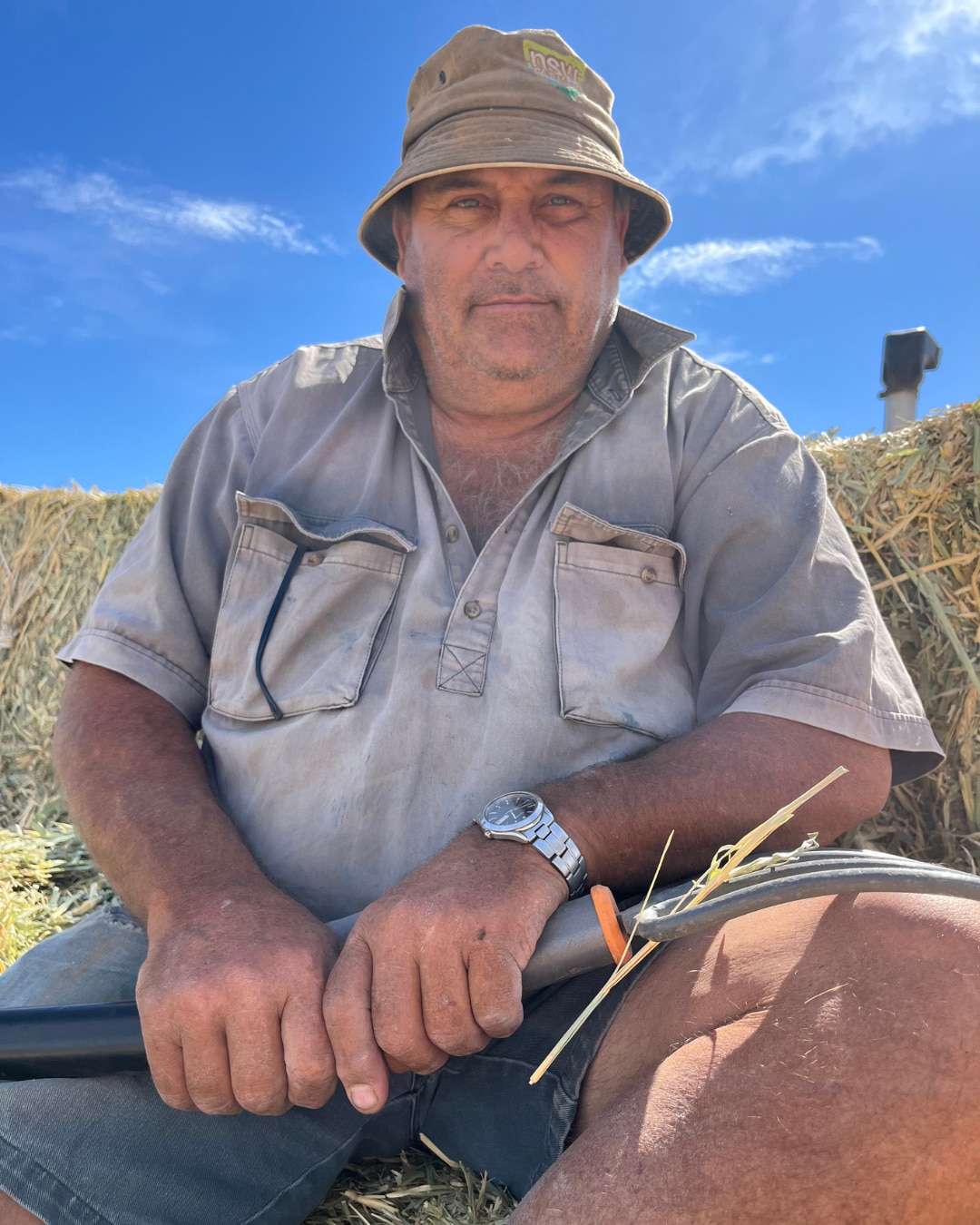 A man in a work shirt and bucket hat sitting on a pile of hay holding secateurs