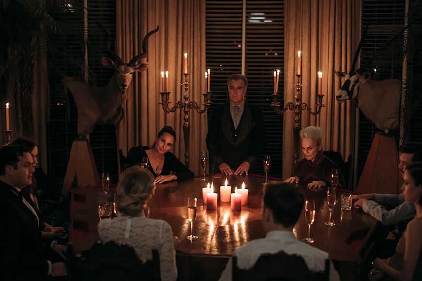Henry Czerny stands between seated Andie MacDowell and Nicky Guadagni at round table in Gothic style room lit up by candles.