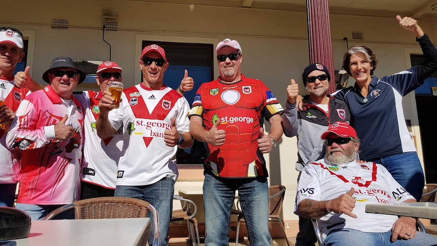 Rugby fans wearing team colours give thumbs at a pub.