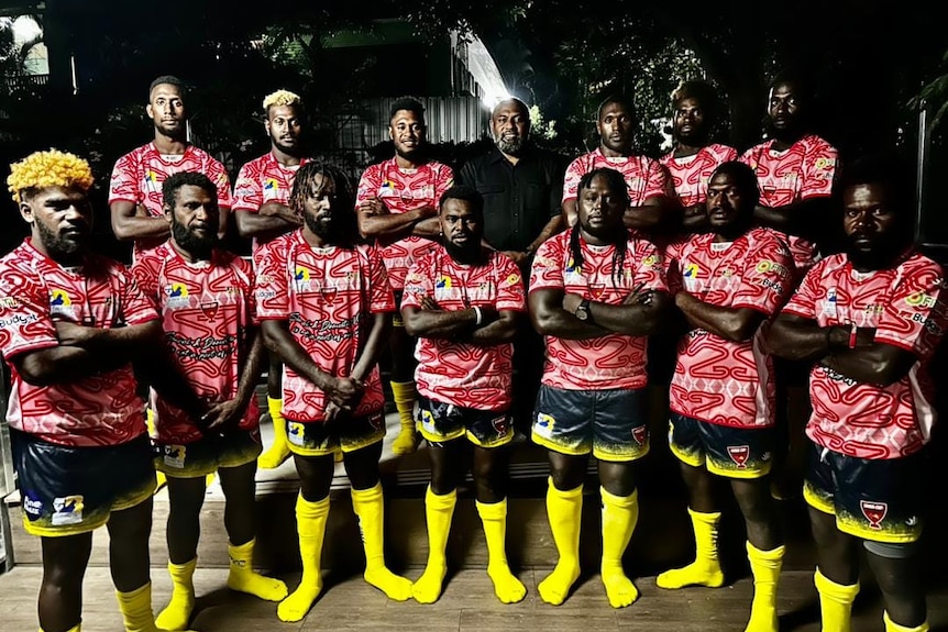Rugby league players in red and white jerseys and yellow shorts stand together, arms folded.