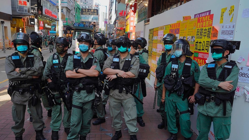 A group of police in riot gear stand on a street, many with arms crossed and truncheons slung from utility vests.