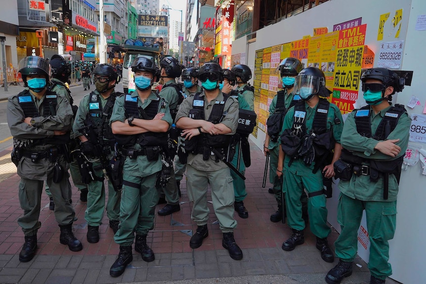 A group of police in riot gear stand on a street, many with arms crossed and truncheons slung from utility vests.