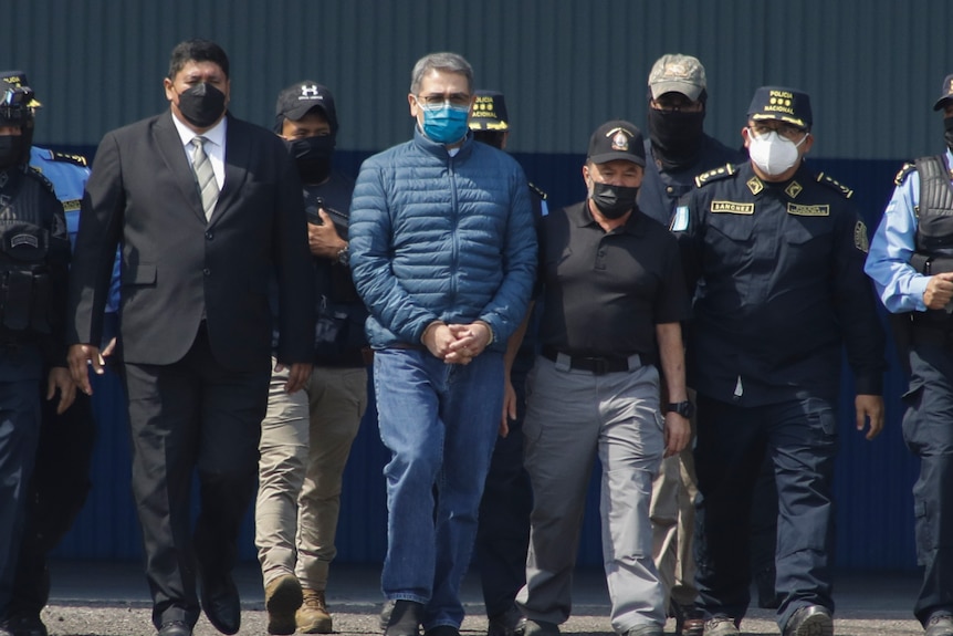 A man in a blue jacket is frog marched by around ten officials in uniform