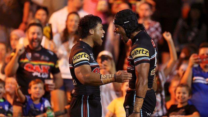 Two NRL players embrace in a hug, in front of a packed grandstand, at night