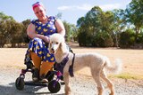 Woman in brightly coloured dress on wheelchair with poodle on gravel trail.