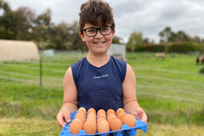 A beaming Fletcher McCulloch stands in front of his paddock, holding a blue tray filled with 30 freshly-laid eggs.