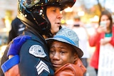 A white police officer waring a helmet embraces a young black boy who has tears running down his face.