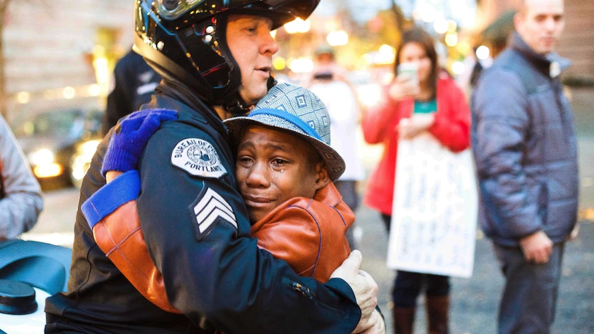 A white police officer waring a helmet embraces a young black boy who has tears running down his face.