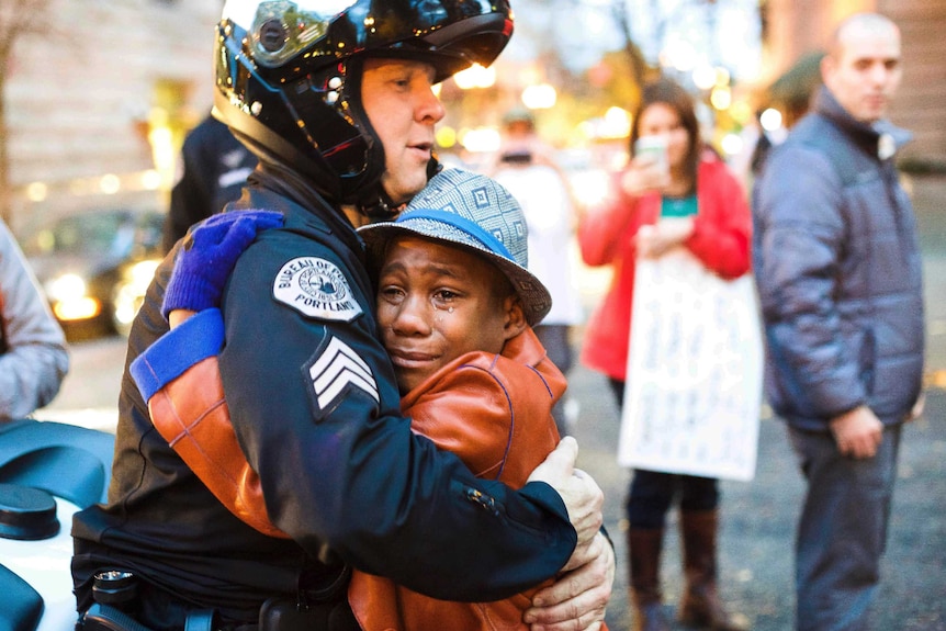 A white police officer waring a helmet embraces a young black boy who has tears running down his face.