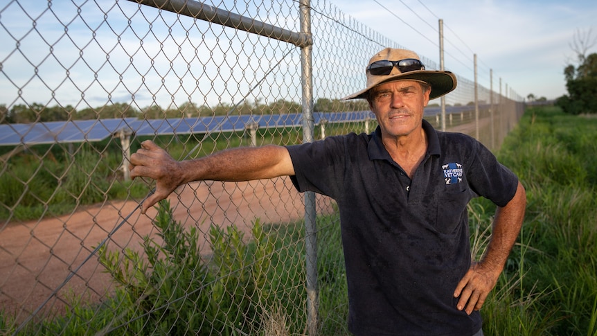 A man leans on a wire fence, with a solar farm in the background. 