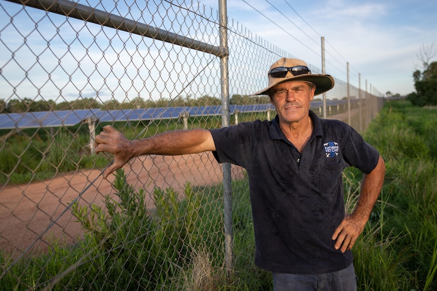A man leans on a wire fence, with a solar farm in the background. 
