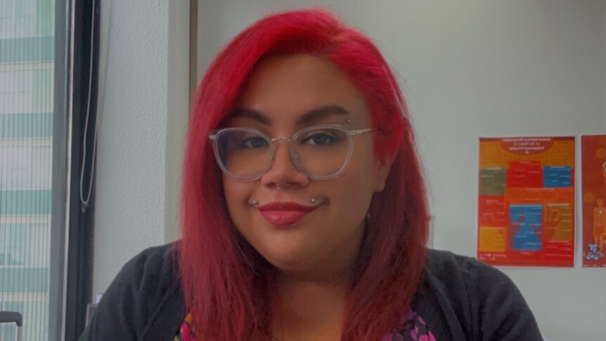 A young woman with bright pink hair and glasses smiles at the camera