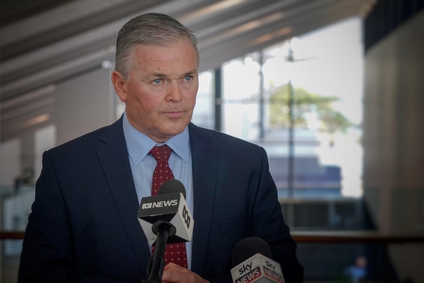 A dark-haired man in a dark suit speaking to the media.