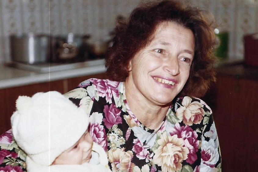 A woman holds a baby in a baptism gown in the kitchen