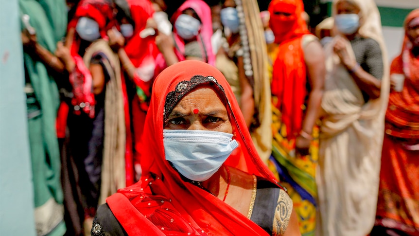 An Indian woman in sari and a surgical face mask looks at the camera with a group of women lined up behind her
