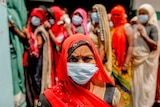 An Indian woman in sari and a surgical face mask looks at the camera with a group of women lined up behind her