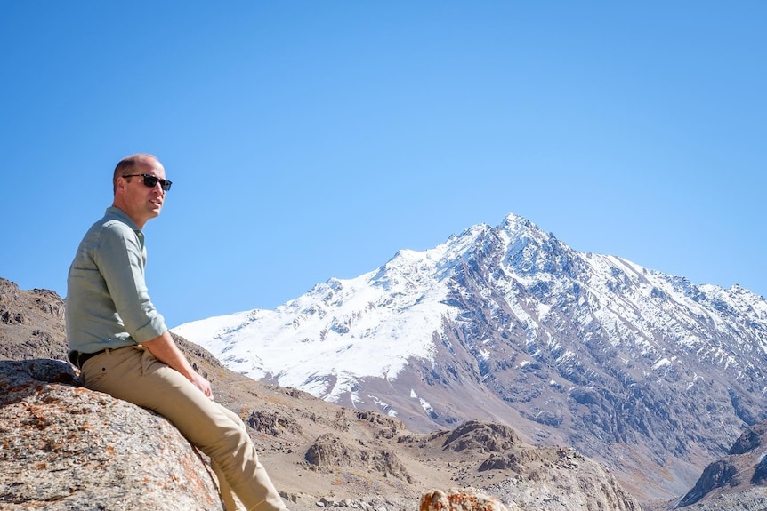 Prince William leans on a rock and looks to the distance. Snow-capped mountaintops can be seen behind him.