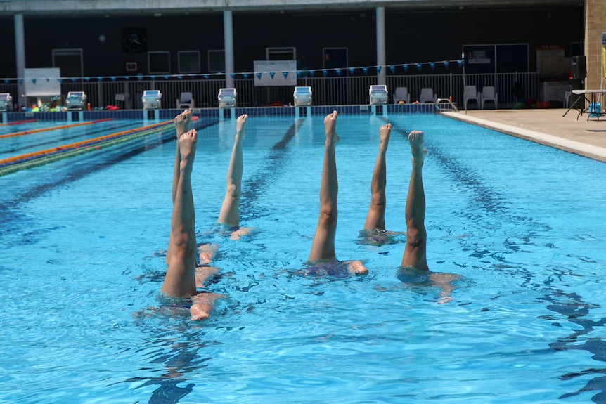 A team of six synchronised swimmers practice in a pool with their legs sticking out above the water.