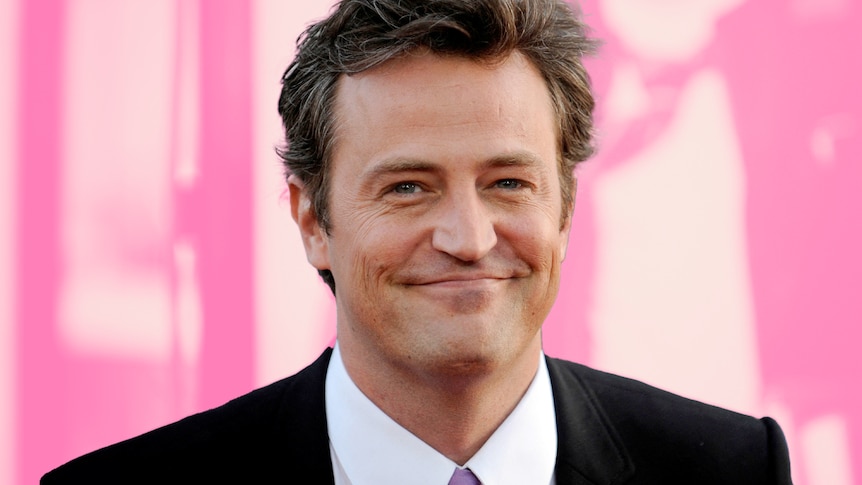 Matthew Perry smiles at camera in front of a pink background