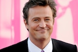 Matthew Perry smiles at camera in front of a pink background