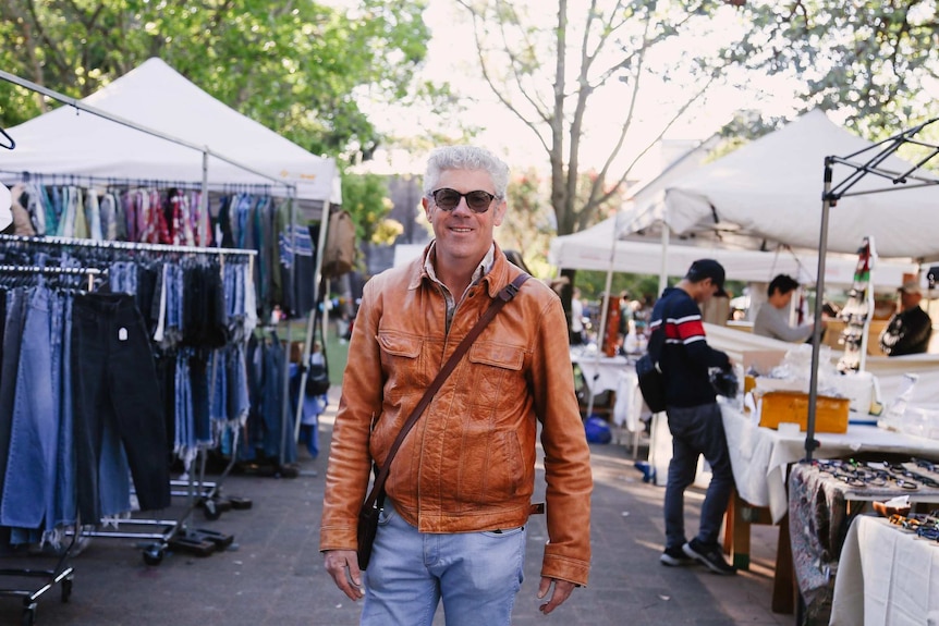 A man in sunglasses and a leather jacket stands in between market stalls