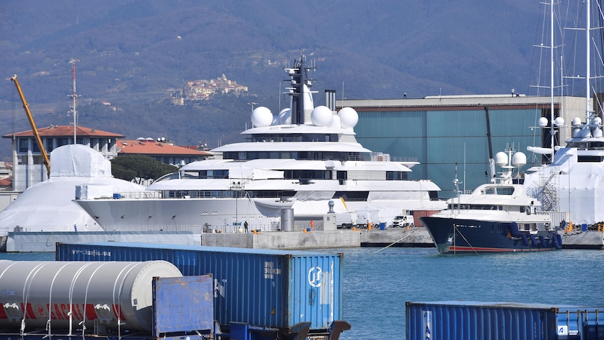 A large superyacht docked in a harbour near smaller boats and shipping containers. Mountainous terrain in the far background.