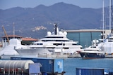 A large superyacht docked in a harbour near smaller boats and shipping containers. Mountainous terrain in the far background.