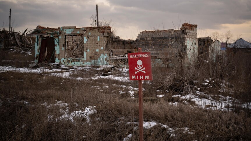 a red sign with a skull and crossbones and reading mines stands in the ground near destroyed buildings