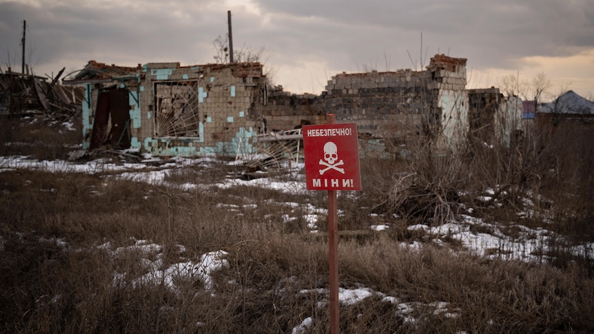 a red sign with a skull and crossbones and reading mines stands in the ground near destroyed buildings