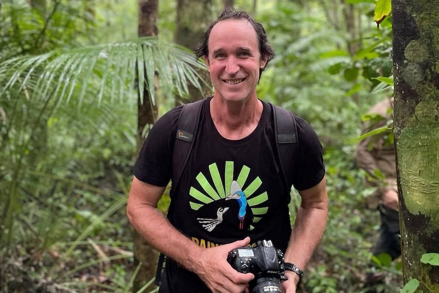 Hombre sonriendo mientras sostiene una cámara en un bosque lluvioso 