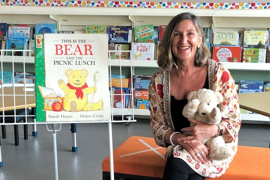 a woman holding a stuffed bear, sitting on a couch in a library with a giant book she is about to read.