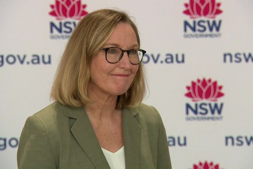 A woman with shoulder-length blonde hair in a bob, with dark-rimmed glasses, green jacket, white shirt in front of NSW backdrop