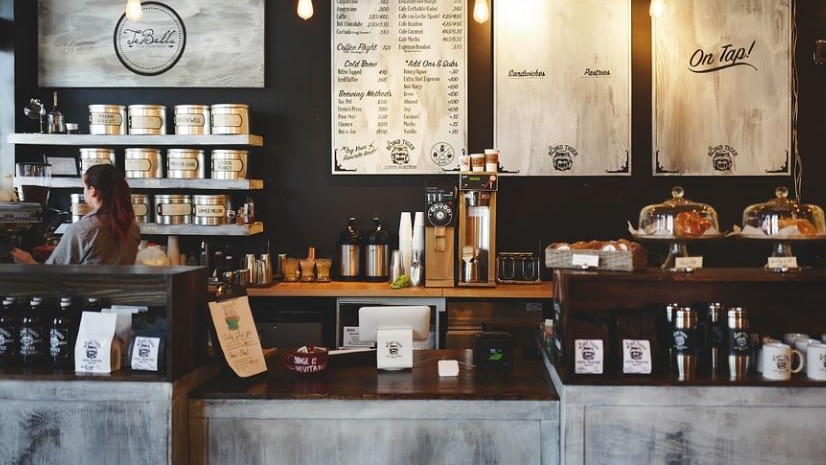 Photo of a cafe behind the counter. There is a woman in the corner with her back turned 