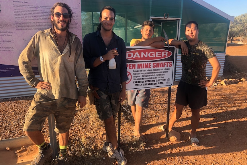 Four men holding opal mining gear stand next to a warning sign.