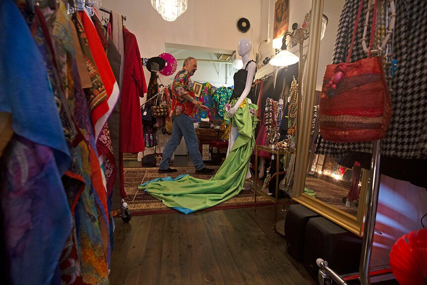 Wide shot of a shop room with a man tending to a mannequin in a black top and elaborate green train shirt.
