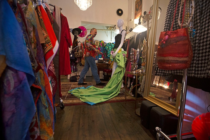 Wide shot of a shop room with a man tending to a mannequin in a black top and elaborate green train shirt.