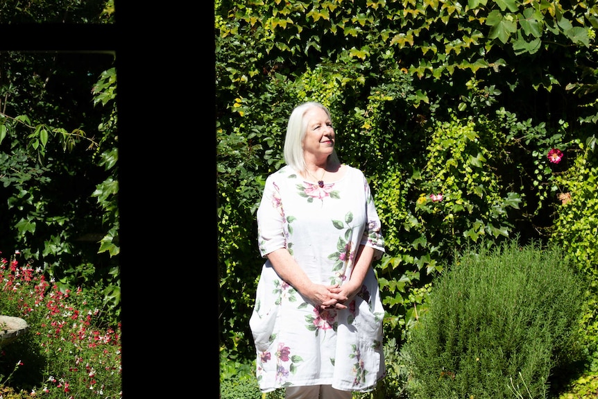 A woman with blonde hair and floral print dress stands looking up smiling, in a garden full of green bushes and vine.