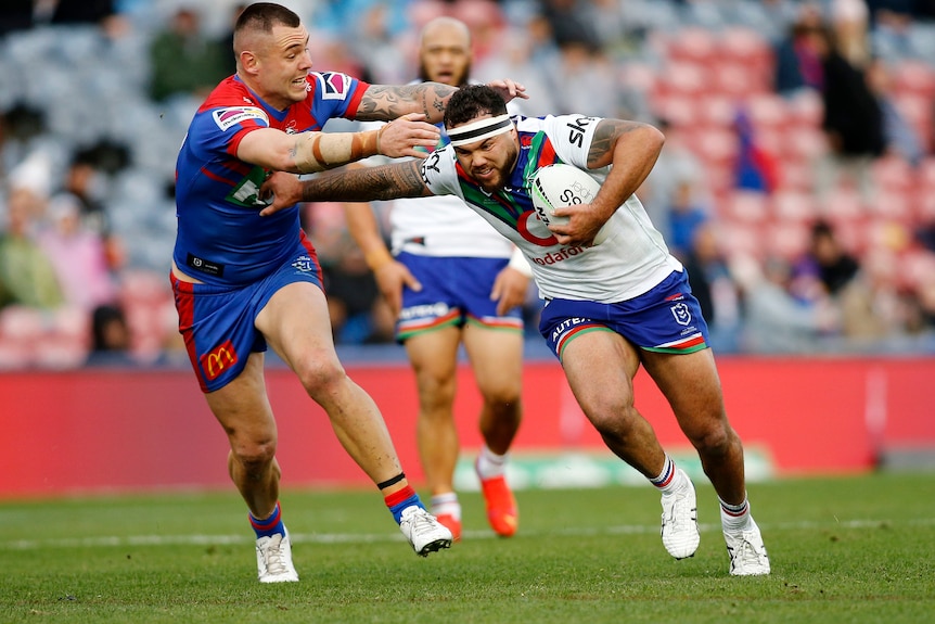 A Warriors NRL player holds the ball with his left hand as he palms off a Newcastle opponent.