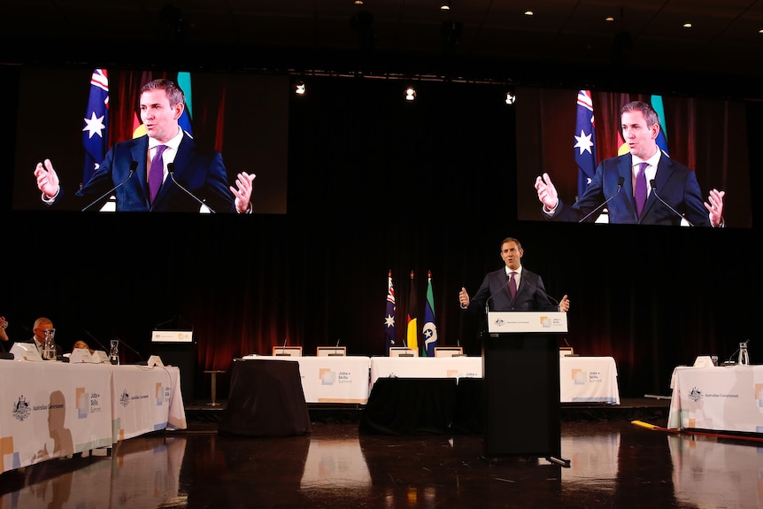 Chalmers speaks from a podium with two large screens displaying his face behind him.