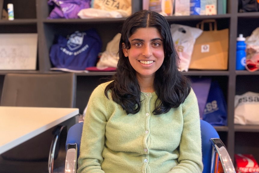 A woman with dark brown hair and a green jumper sitting in a chair and smiling