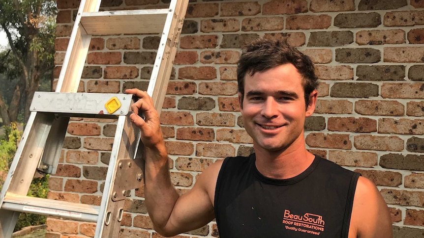 A young man in a black singlet, pictured smiling in front of a brick house. His right hand rests on a ladder.