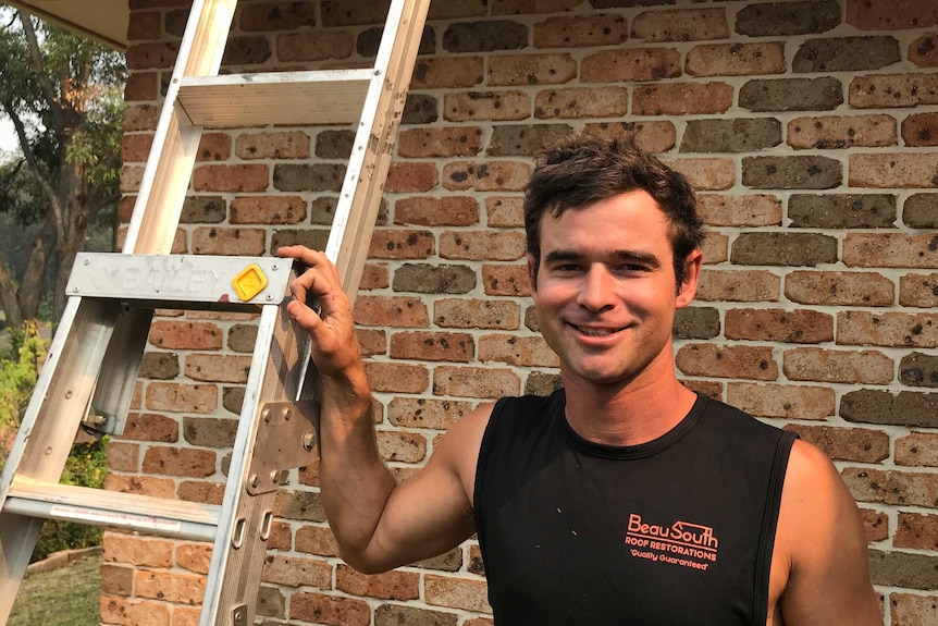 A young man in a black singlet, pictured smiling in front of a brick house. His right hand rests on a ladder.
