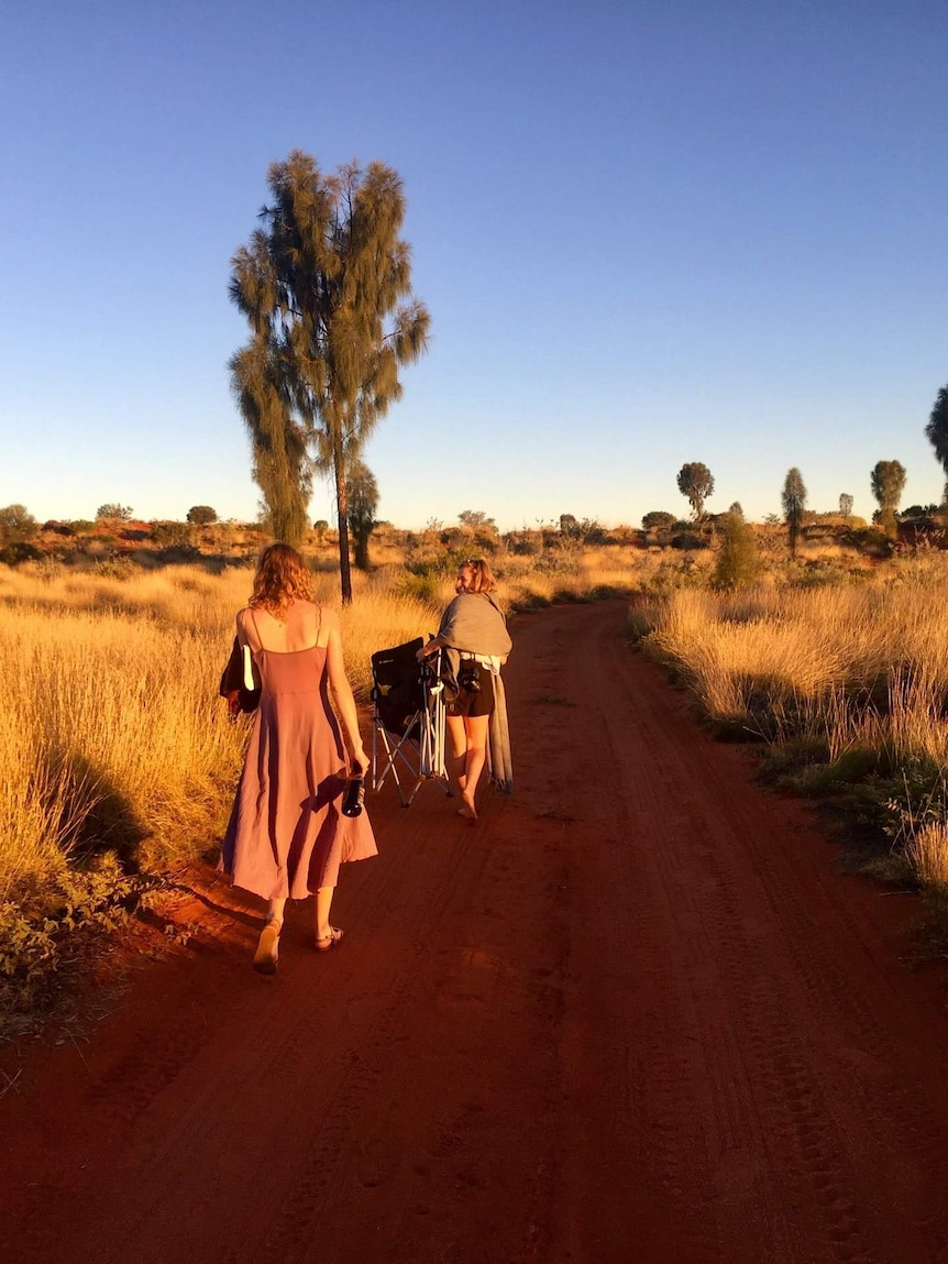 Two women walk along a red dirt track surrounded by spinifex