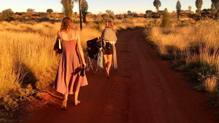 Two women walk along a red dirt track surrounded by spinifex
