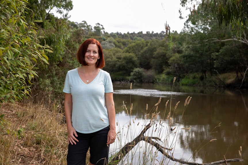 A woman in a blue T-shirt and black pants with red hair cut in a short bob stands beside the Yarra river.