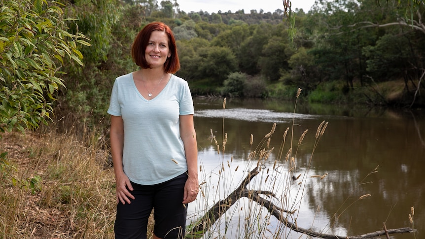A woman in a blue T-shirt and black pants with red hair cut in a short bob stands beside the Yarra river.
