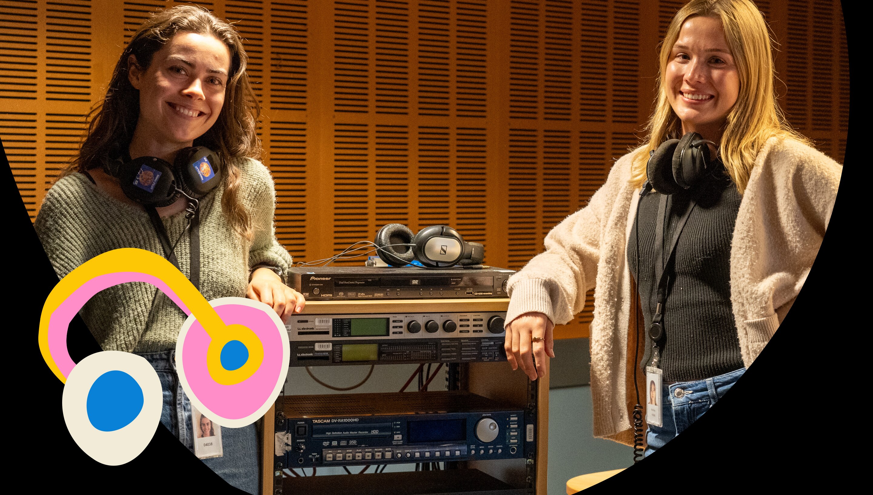 Two young woman in a sound studio wearing headphones. They are smiling and leaning on an audio equipment cabinet.