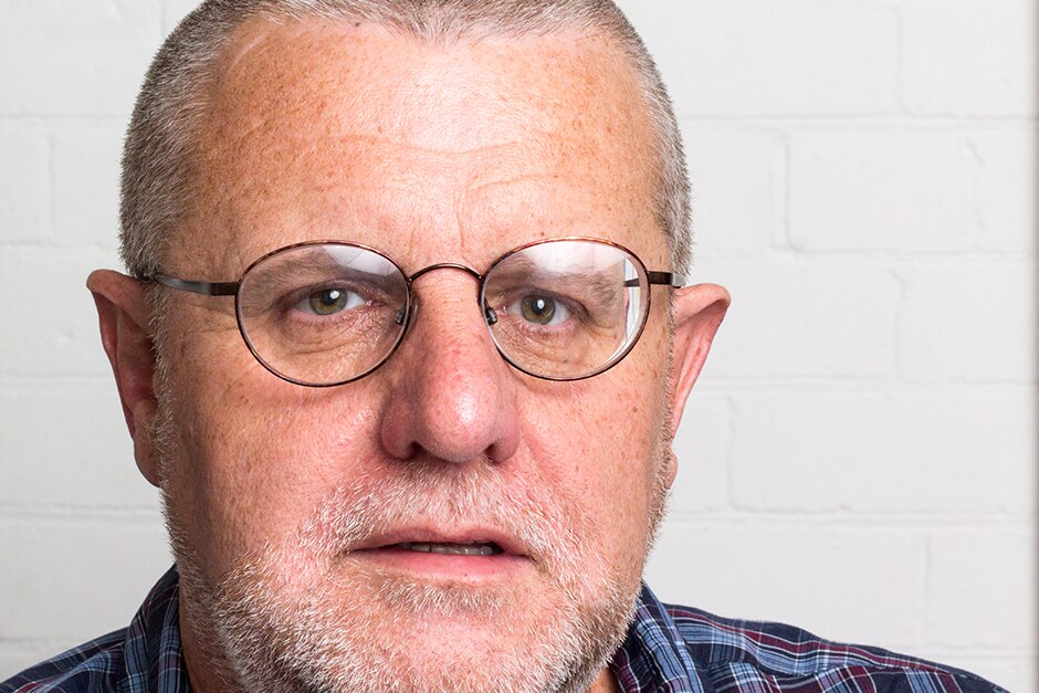 An older man in front of a white wall.