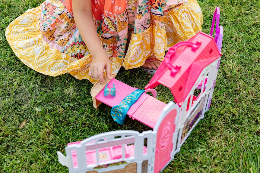 A child plays with a plastic, pink dollhouse on grass.